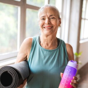 Portrait of an older woman looking at the camera while holding a yoga mat and a water bottle