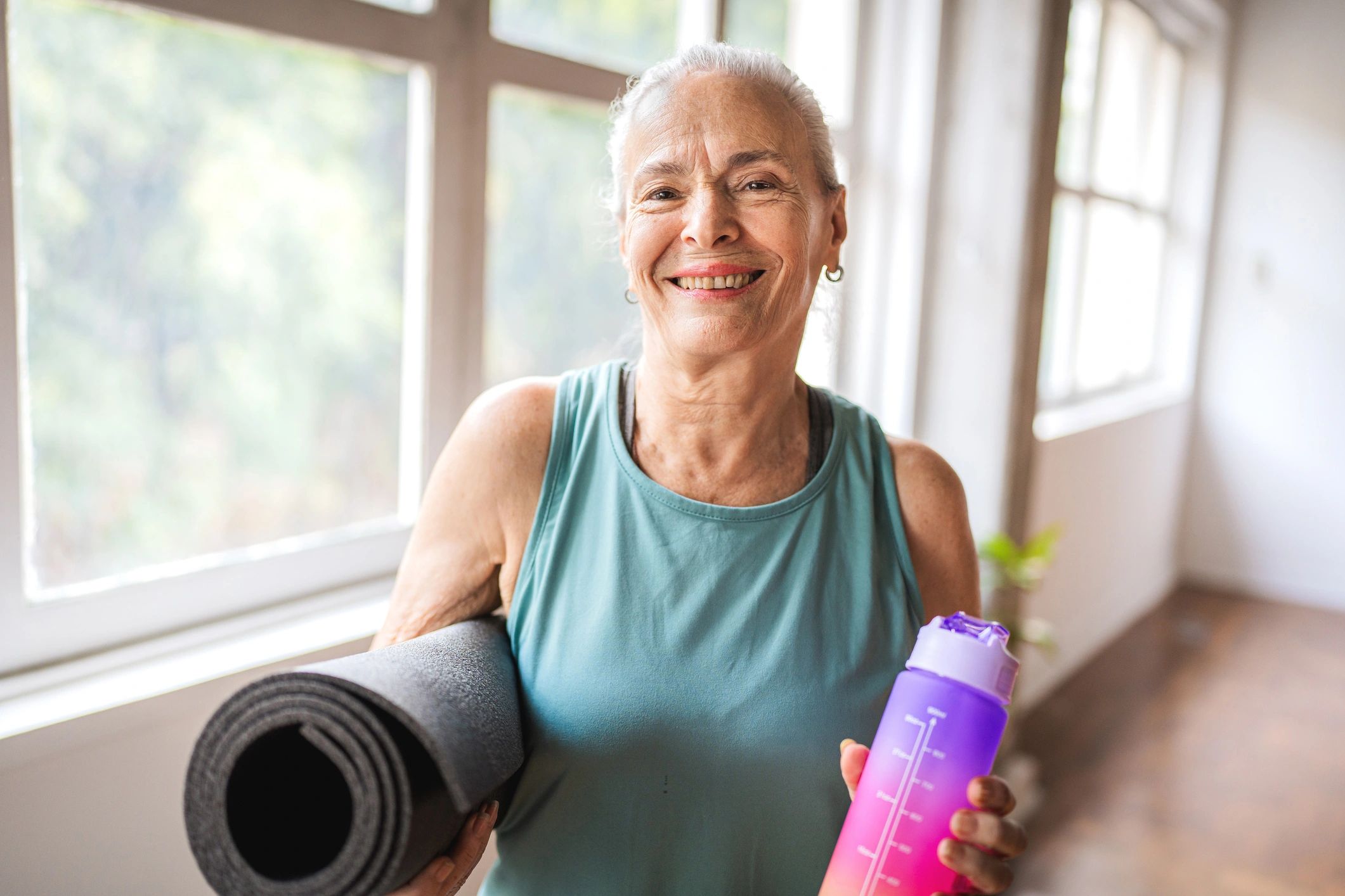 Portrait of an older woman looking at the camera while holding a yoga mat and a water bottle